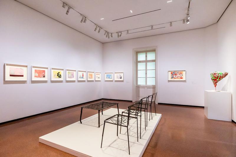 Moderne Galerie mit Kunstwerken an Wänden, einem Tisch und Stühlen in der Mitte sowie einer bunten Skulptur in der Ecke.