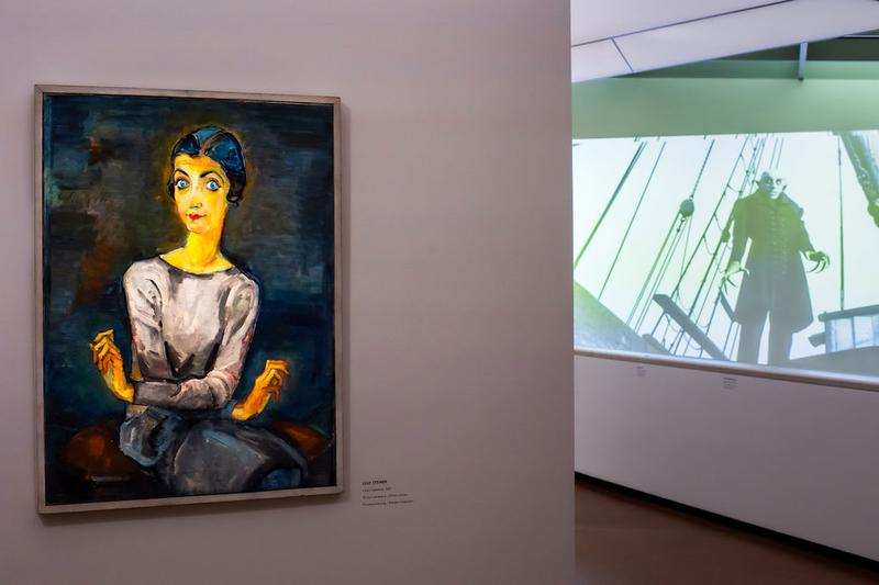 Auf dem Foto sieht man einen Teil der Ausstellung „Experiment Expressionismus“.

Links hängt ein Gemälde von Lilly Steiner.
Es zeigt Lilian Gärtner mit großen, blauen Augen und einem grauen Kleid.
Sie sitzt und hält die Hände in einer besonderen, ausdrucksvollen Haltung.
Das Bild wurde heute schon beschrieben.

Rechts sieht man eine Filmszene aus „Nosferatu“.
Der Film ist schwarz-weiß und wird auf die Wand projiziert.
Man sieht den Vampir Nosferatu auf einem Schiff.
Er steht auf dem Deck und schaut unheimlich.