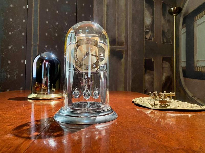 Glasglocke mit einer eleganten Uhr und zwei weiteren Objekten auf einem Holztablett vor einer künstlerischen Wand.