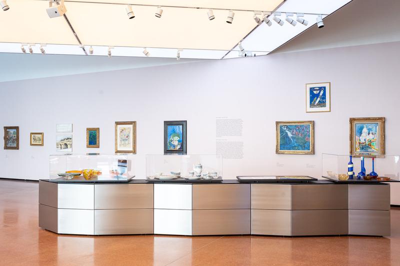 Moderne Galerie mit Gemälden an der Wand und einem Ausstellungsstück aus Glas und Metall in der Mitte.