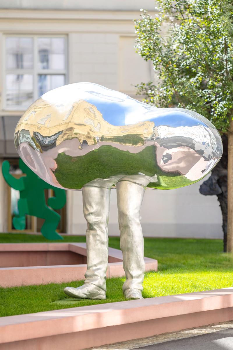 Skulptur mit spiegelndem, bohnenförmigem Oberteil und menschlichen Beinen, umgeben von Grünflächen und weiteren Kunstwerken.