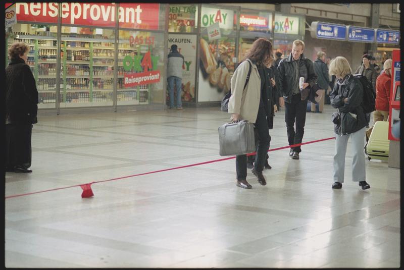 Zu sehen ist die offene Halle des Wiener Westbahnhofs. Im Vordergrund verläuft eine gespannte rote Linie schräg über das Bild – es handelt sich dabei um einen roten Damenstrumpf, der stark in die Länge gezogen wurde. Der Strumpf ist an einem Ende auf dem Boden mit einem kleinen roten Objekt beschwert oder befestigt. Menschen im Bild scheinen sich durch die Situation irritiert, neugierig oder amüsiert zu verhalten. Zwei Frauen im Vordergrund betrachten das Geschehen aus der Nähe: Eine trägt eine helle Jacke und hält eine graue Tasche, die andere trägt eine schwarze Jacke und blonde Haare. Beide stehen dicht an dem gespannten Strumpf. Im Hintergrund gehen weitere Passant:innen vorbei oder bleiben kurz stehen. Links im Bild ist ein Geschäft mit der Aufschrift „Reiseproviant“ zu sehen, daneben befinden sich weitere Läden mit bunten Reklametafeln.

Das Bild stammt aus einem Videoausschnitt einer Performance der Künstlerin Carola Dertnig, die im öffentlichen Raum – hier im Westbahnhof Wien – stattfindet. Der in die Länge gezogene rote Strumpf wird dabei als irritierendes, poetisches Element eingesetzt, das den gewohnten Bewegungsfluss der Passant:innen unterbricht und Fragen nach Raum, Körper, Geschlecht und Aufmerksamkeit aufwirft. Die Künstlerin nutzt alltägliche Materialien und Orte, um performativ neue Bedeutungsräume zu öffnen.