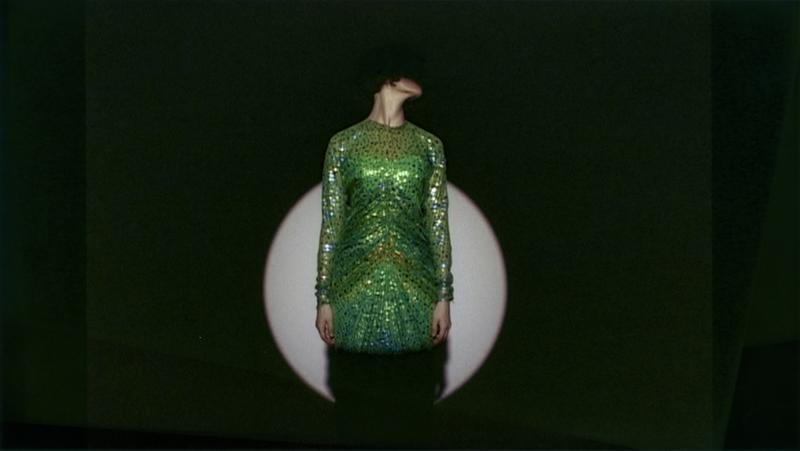 Auf dem Bild sieht man eine Person in einem grünen Kleid.
Das Kleid ist glänzend und mit vielen Pailletten oder Glitzersteinen verziert.
Die Person steht vor einem dunklen Hintergrund,
in dem ein runder, heller Lichtkreis, wahrscheinlich von einem Scheinwerfer, zu sehen ist

Der Kopf der Person ist nach links geneigt,
sodass man das Gesicht nicht sehen kann.
Das macht das Bild geheimnisvoll und spannend.