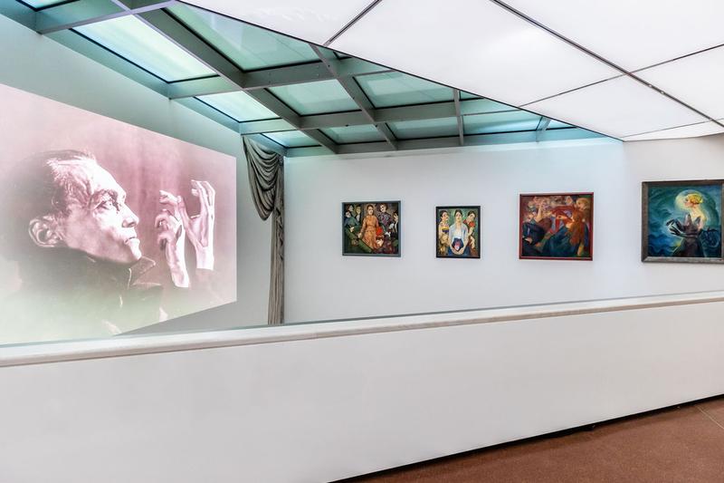 Ganz links sieht man ein Foto , das auf eine Wand projiziert wird. Es zeigt den Schauspieler Conrad Veidt in Schwarz-Weiß.
Er hebt die Hände und schaut ernst.

Rechts daneben hängen vier Gemälde:

– Das erste links zeigt mehrere Menschen. In der Mitte sitzt eine Frau in einem orangefarbenen Kleid.
– Das zweite zeigt drei Frauen.
– Das dritte zeigt Menschen aus der Erzählung Decameron.
– Das vierte ist das Bild „Terzetta“ mit der Frau, die die langen, dunklen Hände hebt.

Die Farben in allen Bildern sind kräftig und leuchtend.