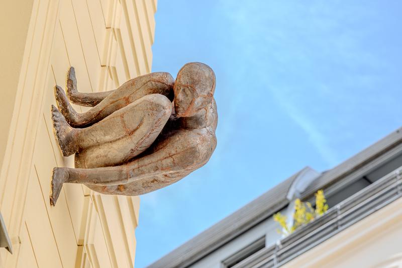 Skulptur eines sitzenden, menschlichen Körpers an einer Wand, vor blauem Himmel und modernen Gebäuden.