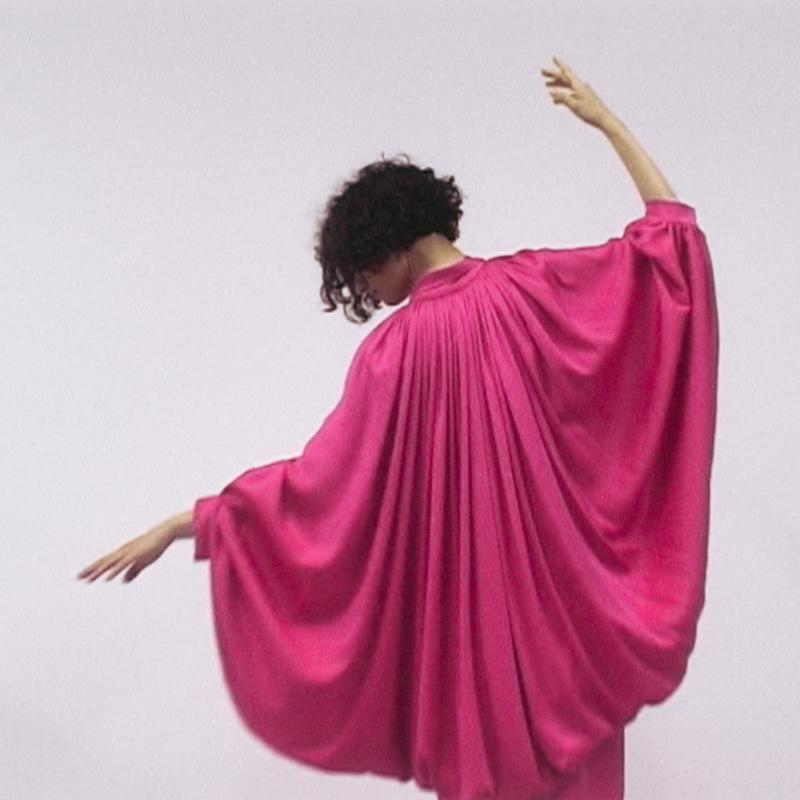 Frau in einem fließenden, pinken Kleid mit weiten Ärmeln, die sich elegant zur Seite dreht.