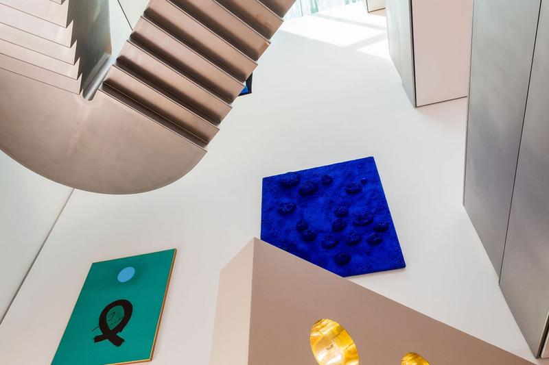 Auf dem Foto sieht man einen Blick nach oben im Museum der Heidi Horten Collection in Wien.
Man erkennt eine moderne Treppe aus Metall und große, helle Wände.

Links an der Wand hängt ein grünes Bild mit einem schwarzen Zeichen und einem blauen Kreis.
Dieses Kunstwerk heißt „Blue Orb“ und ist von Adolph Gottlieb.

Daneben, weiter oben, sieht man ein kräftig blaues Kunstwerk mit Schwämmen darauf.
Das ist das allererste Schwammrelief von Yves Klein.
Er hat echte Schwämme mit tiefblauer Farbe eingefärbt und auf die Leinwand geklebt.
Das Werk ist sehr besonders, weil Yves Klein für diese intensive blaue Farbe berühmt wurde.

Unten im Bild sieht man einen Teil der Bullaugenwand von Markus Schinwald.
Die runden, gold glänzenden Fenster gehören zu einem Gestaltungselement.