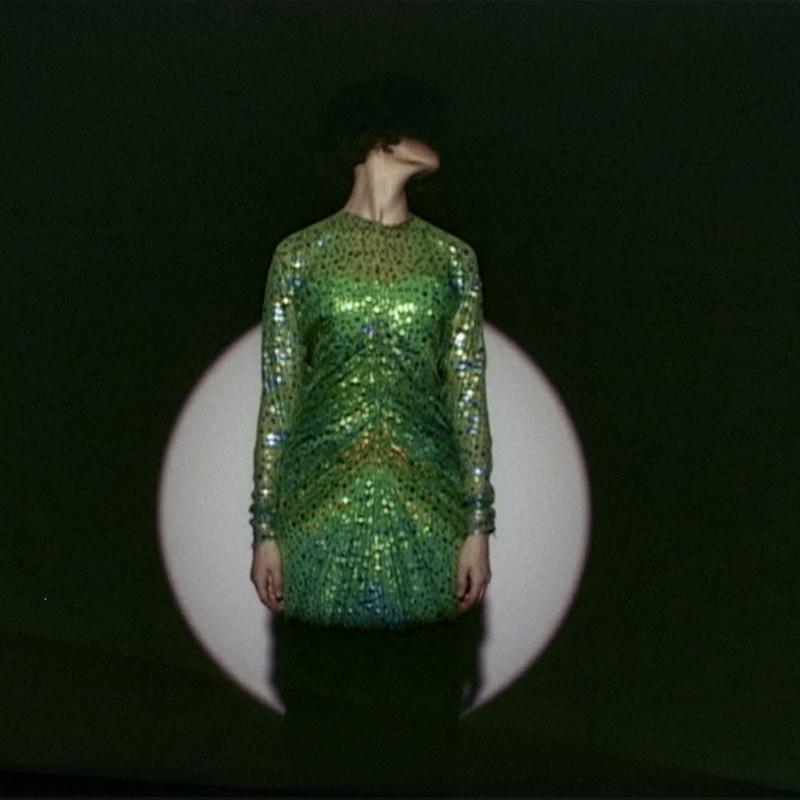 Auf dem Bild sieht man eine Person in einem grünen Kleid.
Das Kleid ist glänzend und mit vielen Pailletten oder Glitzersteinen verziert.
Die Person steht vor einem dunklen Hintergrund,
in dem ein runder, heller Lichtkreis, wahrscheinlich von einem Scheinwerfer, zu sehen ist

Der Kopf der Person ist nach links geneigt,
sodass man das Gesicht nicht sehen kann.
Das macht das Bild geheimnisvoll und spannend.