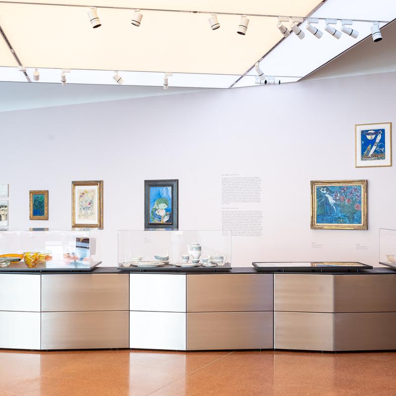 Moderne Galerie mit Gemälden an der Wand und einem Ausstellungsstück aus Glas und Metall in der Mitte.