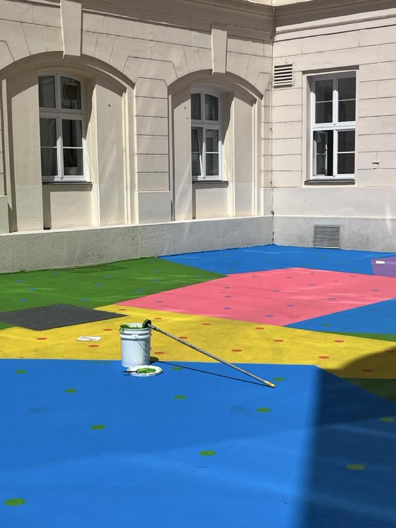 Auf dem Foto sieht man Hanuschhof in Wien. Der Boden ist in starken Farben bemalt – man erkennt große Flächen in Blau, Gelb, Grün, Pink, Schwarz und Lila.

In jeder Farbfläche sind viele kleine, bunte Punkte zu sehen. Diese Punkte zeigen, wie viel CO₂ in einem bestimmten Jahr ausgestoßen wurde – ein Punkt steht für eine Milliarde Tonnen.

Vorne in der Mitte steht ein weißer Farbeimer mit einem langen Stiel, daneben liegt ein kleines Farbtablett – man sieht, dass gerade an dem Kunstwerk gearbeitet wird oder es vor Kurzem fertiggestellt wurde.

Die Farben sind kräftig und leuchten im Sonnenlicht. Die Bemalung macht den Hof freundlich, auffällig und lebendig – und sie hat einen Zweck:
Die Spezialfarbe hilft dabei, die Hitze zu reduzieren, weil sie das Sonnenlicht besser zurückwirft als normaler Beton.

Dieses Kunstwerk heißt „CO₂ntext“. Es verbindet Umweltthema, Kunst und Stadtraum – und lädt zum Nachdenken und Verweilen ein.