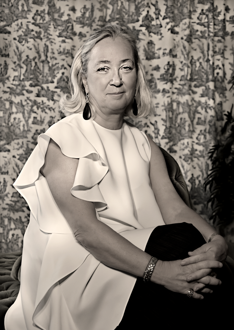 Agnes-Husslein-Arco mit blonden, lockigen Haaren in einem weißen, rüschigen Kleid, sitzend vor einem gemusterten Hintergrund.