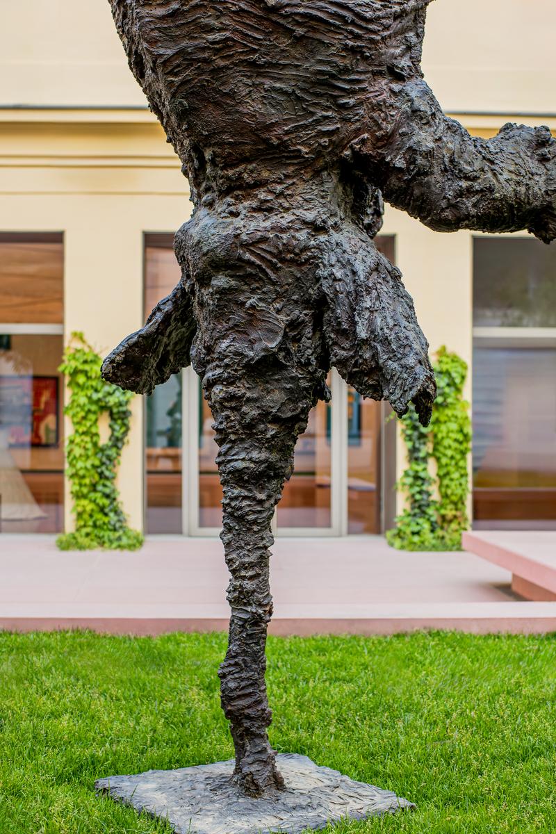Bronzeskulptur eines stehenden Menschen mit texturierter Oberfläche, im Freien vor einem Gebäude mit Pflanzen.