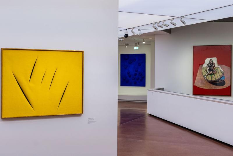 Gelbes Kunstwerk mit Einschnitten, daneben blaue und rote Gemälde in einem modernen Ausstellungsraum.
