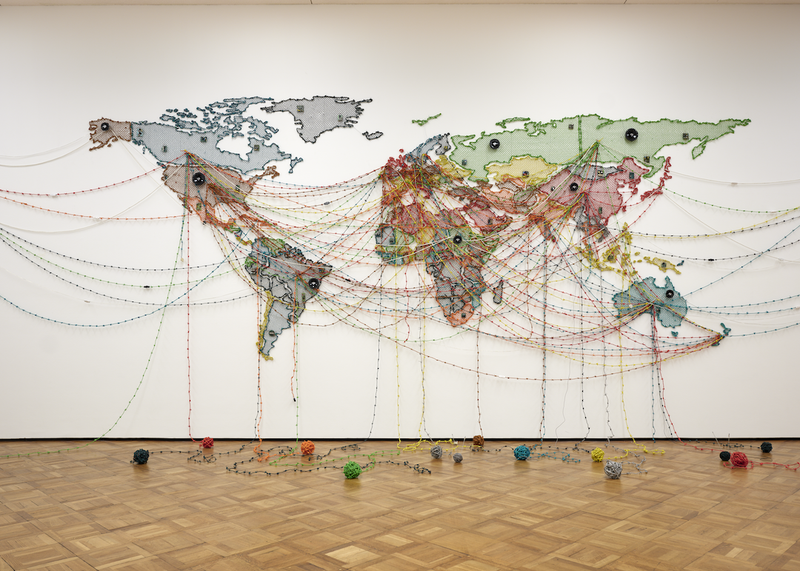 Das Kunstwerk zeigt eine großformatige Weltkarte, die an einer weißen Wand angebracht ist. Die Kontinente sind farbig ausgefüllt, klar voneinander abgegrenzt und bilden eine flache, kartografische Darstellung der Erde. Was die Karte besonders macht, sind die unzähligen bunten Fäden, die über sie gespannt sind. Diese Fäden verlaufen in gebogenen Linien von Kontinent zu Kontinent, kreuzen sich vielfach und verbinden Regionen auf verschiedenen Erdteilen miteinander. Die Fäden bestehen aus textilen Materialien wie Garn oder Kabeln und haben leuchtende Farben – Rot, Blau, Grün, Gelb, Rosa. An den Endpunkten der Linien hängen größere Knäuel, wie Wollbälle, die teils an der Wand befestigt, teils auf dem Boden des Ausstellungsraums verteilt sind. Die Fäden spannen sich über den Raum und verbinden Wand, Werk und Boden visuell miteinander.