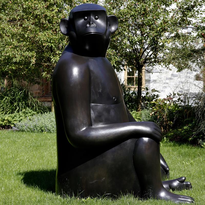 Große, stilisierte Bronze-Skulptur eines sitzenden Affen, umgeben von grünem Gras und Bäumen.