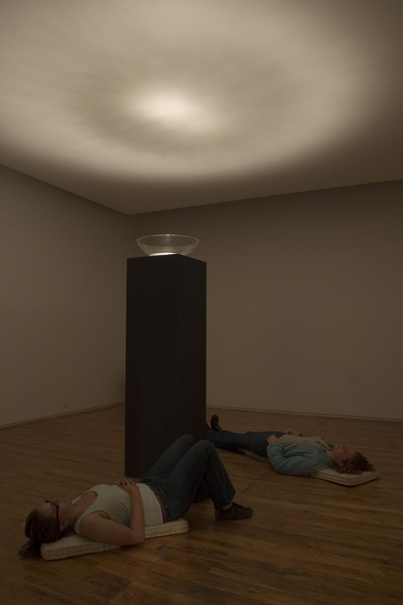 Zwei Personen liegen auf Matten im Raum, während eine Schale auf einem Podest Licht auf die Decke wirft.