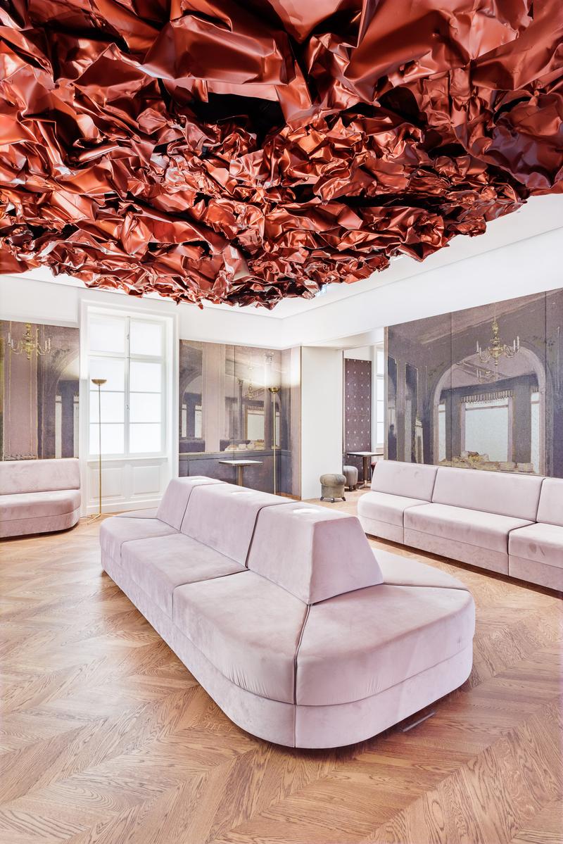 Auf dem Foto sehen wir den Tea Room im Museum der Heidi Horten Collection in Wien.

An der Decke hängt ein großes, rotes Kunstwerk von Hans Kupelwieser.
Es sieht aus wie zerknittertes Papier oder glänzender Stoff,
wurde aber aus Aluminium gemacht.

An der hinteren Wand gibt es viele runde Fenster – sie sehen aus wie Bullaugen.
Dahinter befinden sind kleine, kostbare Objekte aus der Sammlung von Heidi Horten.

Diese Vitrinenwand wurde von Markus Schinwald entworfen.
Auch die rosa Sitzmöbel in der Mitte des Raums gehören zu seinem Konzept.
Sie laden ein, sich hinzusetzen, zu schauen und zu entspannen.

Die Wände sind mit großen Stoffbildern bedeckt,
die an ein altes Café oder Schlosszimmer erinnern.