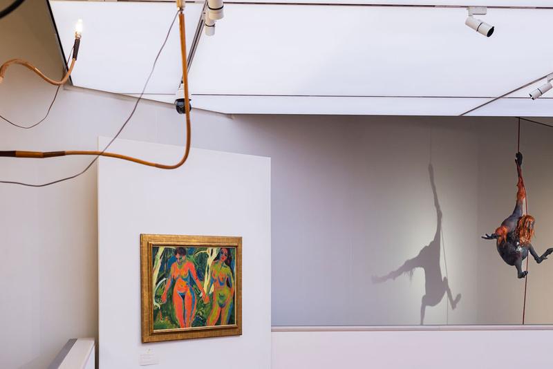 Gemälde mit zwei nackten Frauenfiguren, daneben eine hängende Skulptur mit Schattenwurf in einem modernen Ausstellungsraum.