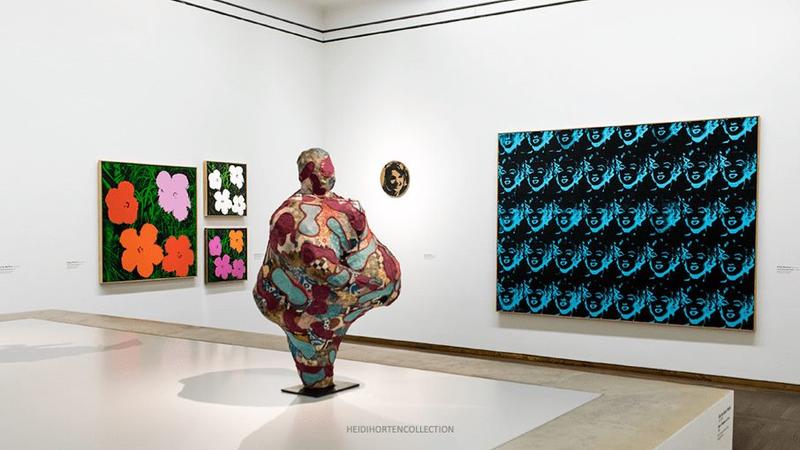 Das Foto zeigt einen hellen, modernen Ausstellungsraum Leopold. Im Mittelpunkt steht eine große, farbenfrohe Skulptur von Niki de Saint-Phalle. An den Wänden sieht man Werke von Andy Warhol. Seine Farbenfrohen Blumen, ein Druck mit Marylin Monroe und ein kleines rundes Werk, wo Jacky Kennedy abgebildet ist. Der Raum ist klar und ruhig gestaltet, mit weißem Boden und hohen Decken.