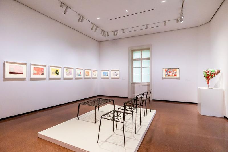 Ausstellungsraum mit bunten Zeichnungen an der Wand, einem Tisch und Stühlen in der Mitte sowie einer Skulptur in der Ecke.