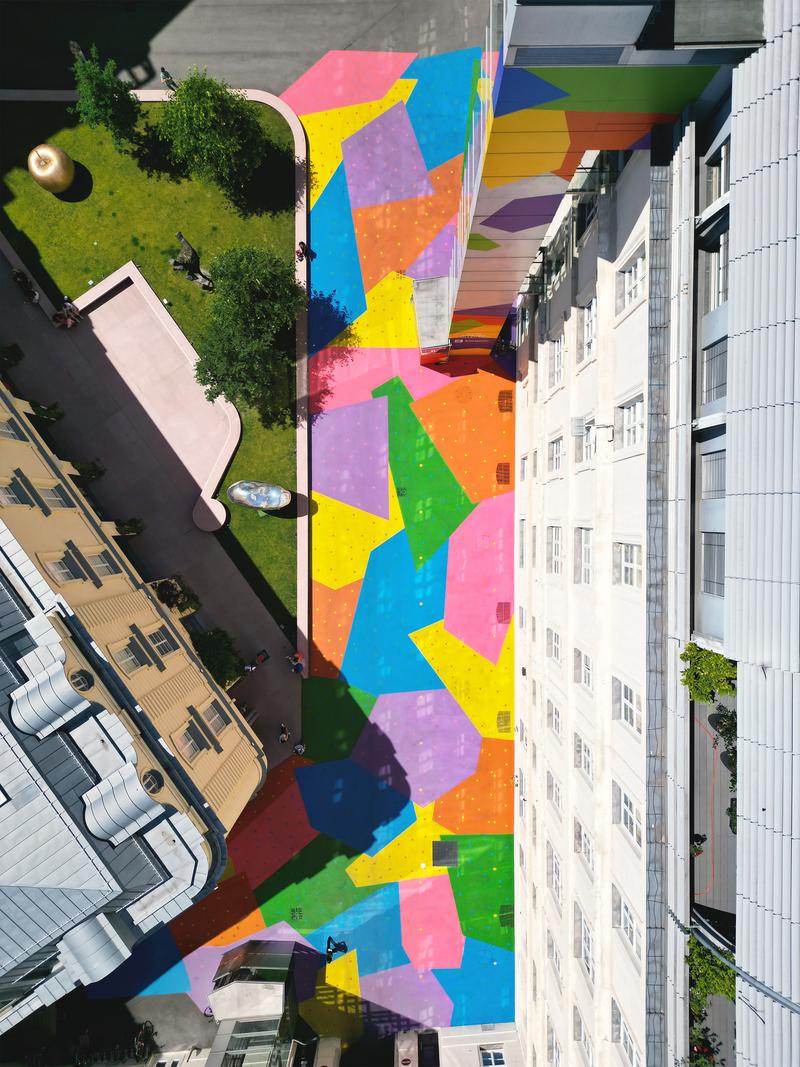 Auf dem Foto sieht man den Hanuschhof in Wien von oben. Die Häuser im Hof sind hell und der Boden und Lift sind in starken Farben bemalt – man erkennt große Flächen in Blau, Gelb, Grün, Pink, Schwarz und Lila.

In jeder Farbfläche sind viele kleine, bunte Punkte zu sehen. Diese Punkte zeigen, wie viel CO₂ in einem bestimmten Jahr ausgestoßen wurde – ein Punkt steht für eine Milliarde Tonnen.

Die Farben sind kräftig und leuchten im Sonnenlicht. Die Bemalung macht den Hof freundlich, auffällig und lebendig – und sie hat einen Zweck:
Die Spezialfarbe hilft dabei, die Hitze zu reduzieren, weil sie das Sonnenlicht besser zurückwirft als normaler Beton.

Dieses Kunstwerk heißt „CO₂ntext“. Es verbindet Umweltthema, Kunst und Stadtraum – und lädt zum Nachdenken und Verweilen ein.

Links oben sieht man einen Skulpturengarten von oben.