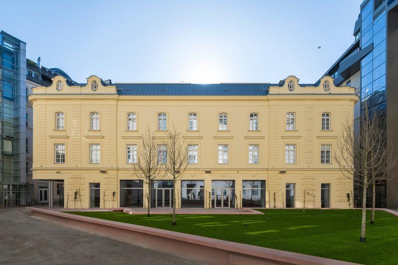 Historisches, gelbes Gebäude mit großen Fenstern, umgeben von modernem Glasbau und grünem Rasen.