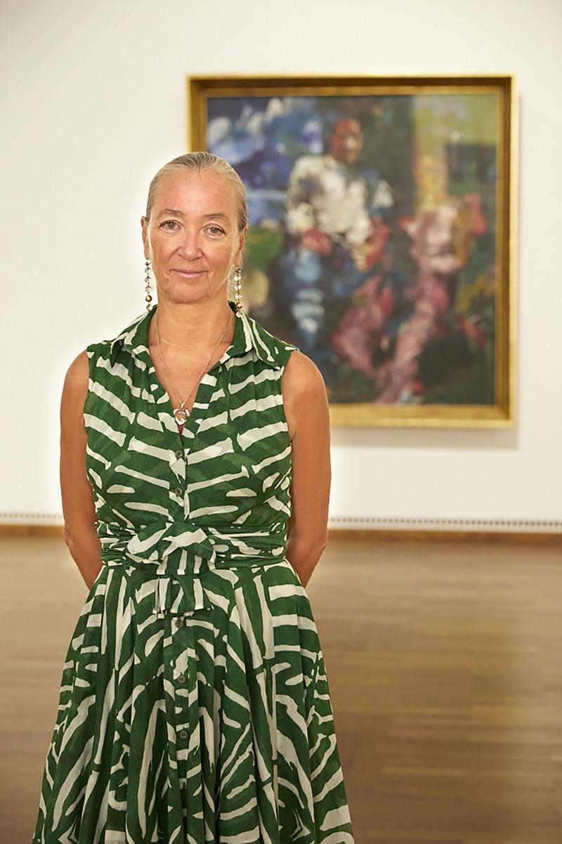 Agnes Husslein-Arco mit langen, blonden Haaren in grün-weiß gestreiftem Kleid vor einem Gemälde in einer Galerie.