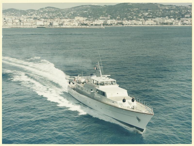 Luxusyacht fährt mit Schaumkronen durch das Wasser, im Hintergrund eine Küstenstadt und Hügel.