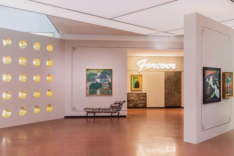 Auf dem Foto sieht man einen Teil der Ausstellung "Klimt⇄Warhol" im Museum der Heidi Horten Collection in Wien.

Links sieht man eine Wand mit runden Fenstern, die wie Bullaugen aussehen.
Diese Wand wurde von dem Künstler Markus Schinwald entworfen.

In der Mitte, direkt vor uns, hängt ein Gemälde von Gustav Klimt.
Darauf sieht man den Attersee, einen See in Österreich.
Das Bild zeigt Wasser, Häuser und Natur in hellen Farben.

Die Verzierungen an den Wänden und die gemalten Möbelstücke neben und unter den Kunstwerken hat ebenfalls Markus Schinwald gestaltet.

Hinten an der Wand, über einem Bild, leuchtet in großen weißen Buchstaben das Wort „Forever“.
Diese Leuchtschrift ist ein Kunstwerk von Tim Noble & Sue Webster.
Darunter hängt ein Selbstporträt von Edvard Munch.

Ganz rechts ragt eine weitere Wand schräg in den Raum hinein.
Dort hängen zwei Gemälde von Emil Nolde, die ebenfalls Teil der Sammlung sind.