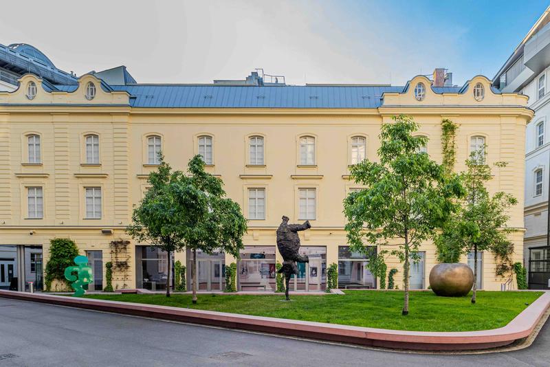 Auf dem Foto sieht man das Gebäude der Heidi Horten Collection in Wien.
Es ist ein hellgelbes Haus mit neoklassizistischer Fassade und großen Fenstern.
Das Gebäude steht im Hanuschhof, einem ruhigen Innenhof mitten in der Stadt.

Vor dem Museum ist ein kleiner Skulpturengarten mit Gras, Bäumen und Kunstwerken.
Man sieht dort drei verschiedene Skulpturen:

Ganz links steht eine grüne Figur von Keith Haring.

In der Mitte steht eine dunkle Bronzefigur, ein Elefant von Miquel Barceló.

Ganz rechts sieht man ein großer goldener Apfel. Er stammt von Claude Lalanne.