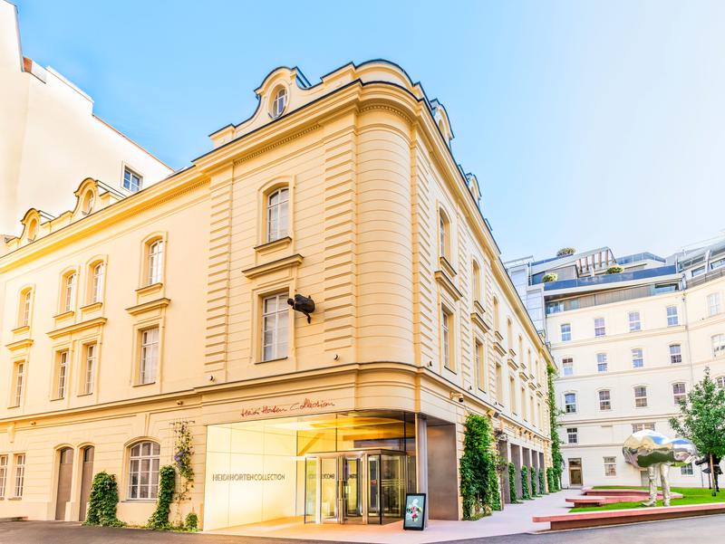 Elegantes Gebäude mit gelber Fassade, Glas-Eingang und grüner Bepflanzung, umgeben von modernen Strukturen.