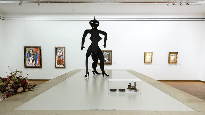 Schwarze Skulptur in der Mitte, umgeben von Gemälden an der Wand und einer Vitrine mit Kunstobjekten.