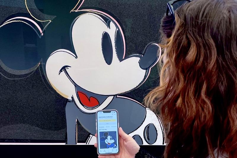 Auf dem Foto sieht man eine Frau von hinten, die sich ein Kunstwerk von Andy Warhol anschaut. Auf dem Bild ist Micky Maus. Die Frau hält ein Smartphone in der Hand. Darauf ist die App Smartify geöffnet – ein Audioguide, der Informationen über Kunstwerke gibt. Man sieht, dass sie genau das Bild auf dem Handy geöffnet hat, das auch vor ihr an der Wand hängt. Das Foto zeigt, wie digitale Medien im Museum helfen, mehr über Kunst zu erfahren – einfach und direkt am eigenen Handy