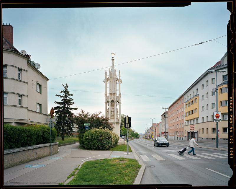 Auf dem Foto sehen wir die Spinnerin am Kreuz.
Das ist ein hoher steinerner Turm mit feinen Verzierungen.
Er steht in Wien, zwischen einer Straße und einem Gehweg.

Links und rechts vom Turm sieht man Wohnhäuser.
Ein Auto fährt auf der Straße.
Ein Mensch geht gerade mit einem Koffer über den Zebrastreifen.

Der Himmel ist blau mit leichten Wolken.
Man sieht viele Details aus dem Stadtleben, zum Beispiel Straßenschilder, Gehsteige, Häuser und Grünflächen.

Das Foto ist mit einer analogen Kamera gemacht, man sieht es an dem schwarzen Filmrand mit dem Schriftzug „Kodak Portra 400“.
Der Fotograf heißt Stefan Oláh.