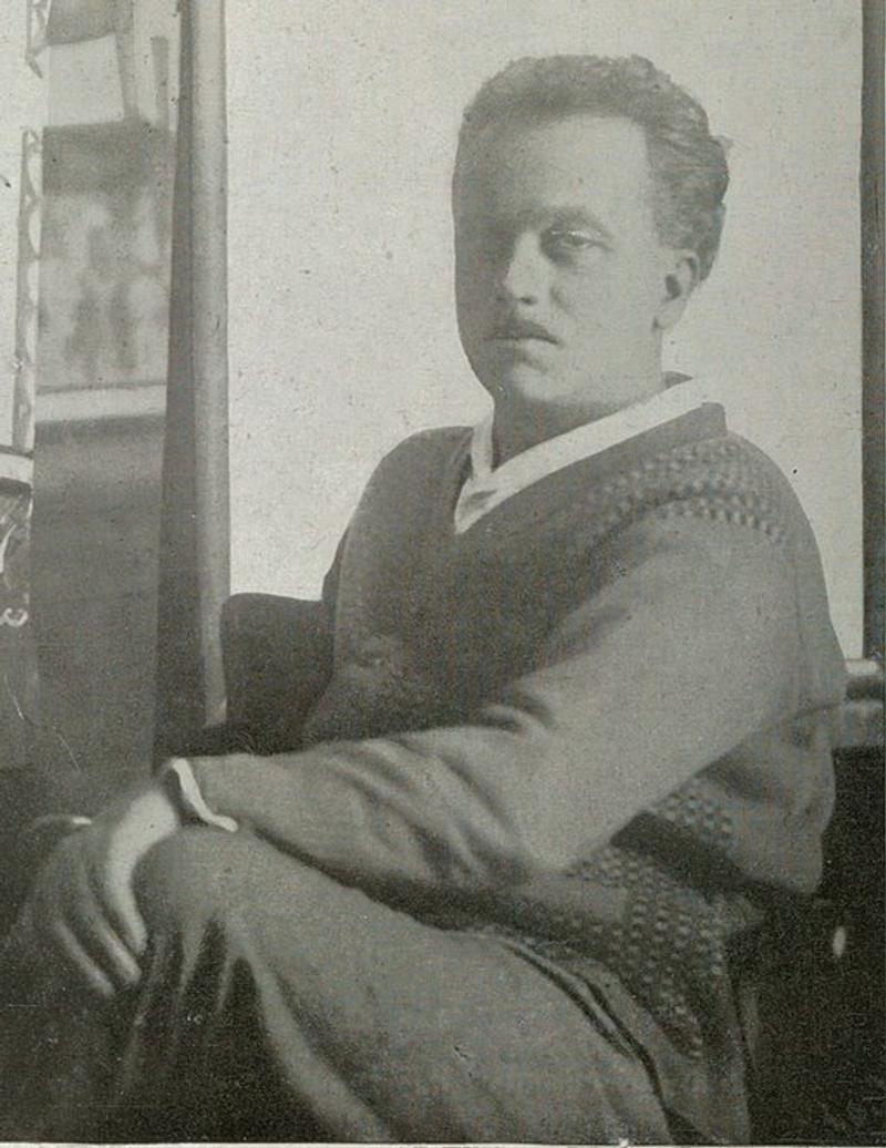 Raoul Dufy mit kurzem Haar, in einem Pullover sitzend, Blick zur Seite, im Hintergrund unscharfe Kunstwerke.