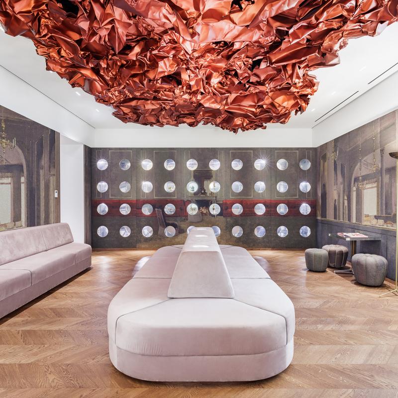 Auf dem Foto sehen wir den Tea Room im Museum der Heidi Horten Collection in Wien.

An der Decke hängt ein großes, rotes Kunstwerk von Hans Kupelwieser.
Es sieht aus wie zerknittertes Papier oder glänzender Stoff,
wurde aber aus Aluminium gemacht.

An der hinteren Wand gibt es viele runde Fenster – sie sehen aus wie Bullaugen.
Dahinter befinden sind kleine, kostbare Objekte aus der Sammlung von Heidi Horten.

Diese Vitrinenwand wurde von Markus Schinwald entworfen.
Auch die rosa Sitzmöbel in der Mitte des Raums gehören zu seinem Konzept.
Sie laden ein, sich hinzusetzen, zu schauen und zu entspannen.

Die Wände sind mit großen Stoffbildern bedeckt,
die an ein altes Café oder Schlosszimmer erinnern.