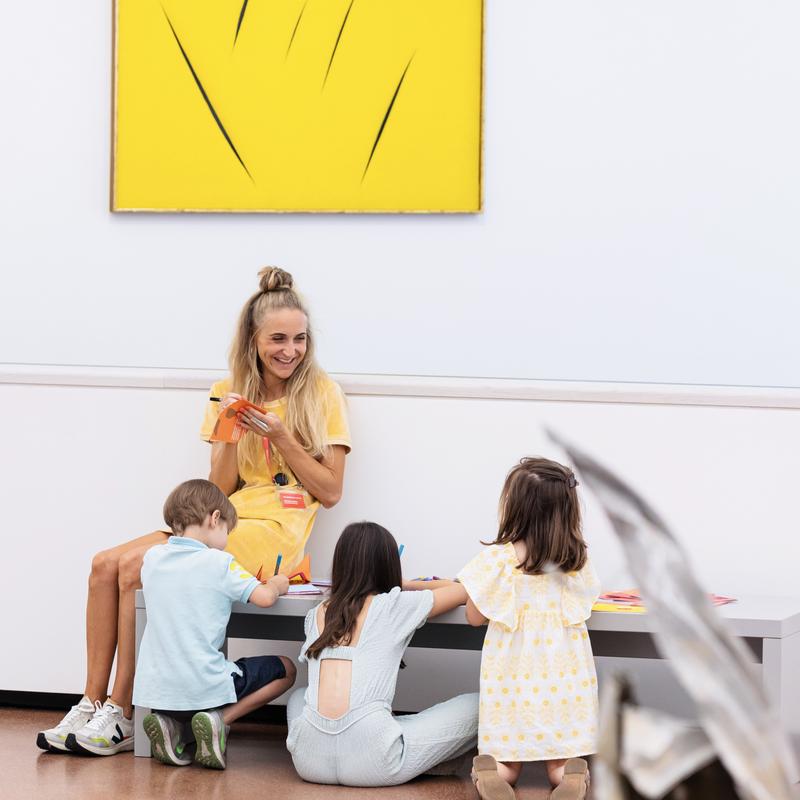 Auf dem Bild sieht man eine Kunstvermittlerin und drei Kinder, die gemeinsam an einem kleinen Tisch im Museum basteln.

Die Vermittlerin trägt ein gelbes Kleid und lächelt, während sie mit buntem Papier arbeitet.
Die Kinder schneiden, kleben oder malen – sie sind konzentriert und mit Freude dabei.

Im Hintergrund hängt ein gelbes Kunstwerk mit schwarzen Rissen an der Wand. Es ist von Lucio Fontana.
Der Raum ist hell und freundlich.

Das Foto zeigt eine kreative Aktion im Rahmen eines Kunstvermittlungsprogramms für Kinder in der Heidi Horten Collection.