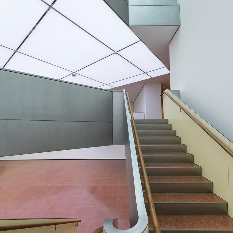 Moderne Treppe mit Holzgeländer, helle Wände und geometrische Decke in einem lichtdurchfluteten Raum.