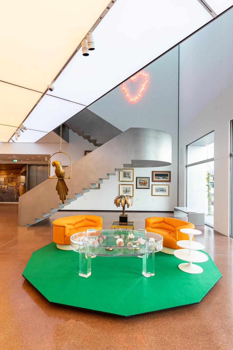 Moderne Lounge mit orangefarbenen Sesseln, gläsernem Tisch, grüner Teppich und einer Treppe im Hintergrund.