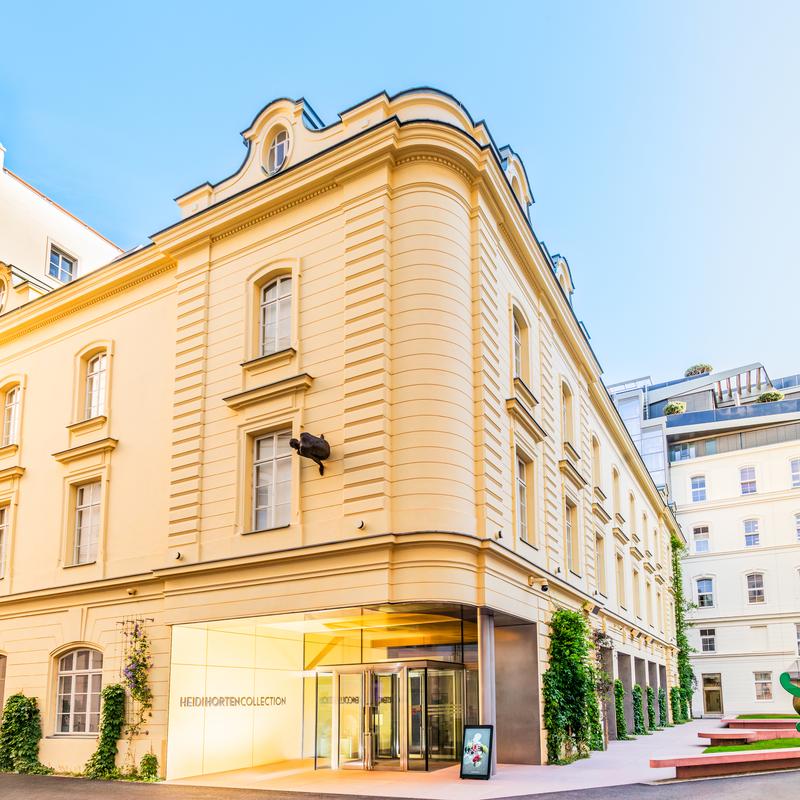 Auf dem Bild sieht man das Gebäude der Heidi Horten Collection.
Es steht im Hanuschhof, im Zentrum von Wien.

Das Haus ist hellgelb und hat eine neoklassizistische Fassade mit großen Fenstern.
Die Form erinnert an ein altes Stadtpalais, wurde aber modern renoviert.

Am Eingang ist eine große Glastür.
Daneben steht in silberner Schrift: „Heidi Horten Collection“.
Ein digitaler Bildschirm zeigt Informationen über das Museum.

Rechts im Bild sieht man einen kleinen Garten mit Skulpturen.
Eine große grüne Figur ist von dem Künstler Keith Haring.

Das Foto zeigt das Museum bei schönem Wetter.
Der Himmel ist blau und das Licht macht den Ort freundlich und einladend.
