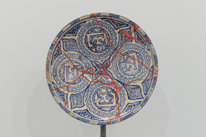 Diese Arbeit von Kader Attia zeigt eine zerbrochene Keramikschale, die sichtbar und bewusst mit leuchtend rotem Harz repariert wurde. Die ornamentalen blauen Muster und kalligrafischen Elemente auf der Oberfläche verweisen auf die kunsthandwerklichen Traditionen der islamischen Welt, insbesondere auf Keramiken aus Nordafrika oder dem Nahen Osten. Statt die Bruchstellen zu verbergen, hebt der Künstler sie deutlich hervor und macht sie zum zentralen Bestandteil der Ästhetik. In Anlehnung an die japanische Kintsugi-Technik, jedoch mit politischer Schärfe, versteht Attia Reparatur nicht als Wiederherstellung eines ursprünglichen Zustands, sondern als einen Akt der Würdigung von Verletzung, Geschichte und Transformation. Die sichtbar gemachten Brüche erzählen von Zerstörung und Heilung – und stehen sinnbildlich für kulturelle Identitäten, die durch Kolonialismus und Migration geprägt und verletzt wurden. Die Schale wird so zu einem stillen Zeugnis von Verlust und Widerstand, aber auch von Resilienz und Neubeginn.