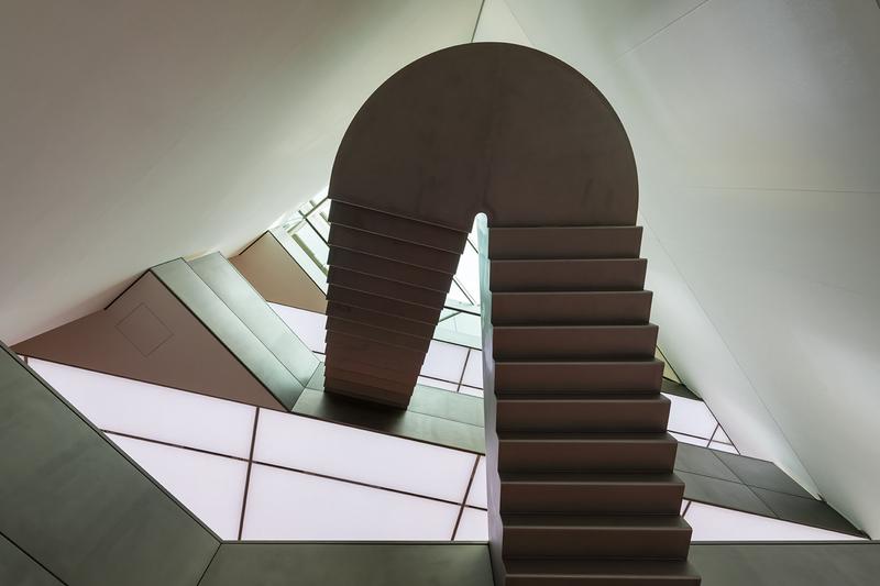 Schwebende Treppe mit geschwungener Form, umgeben von geometrischen Wänden und sanftem Licht.