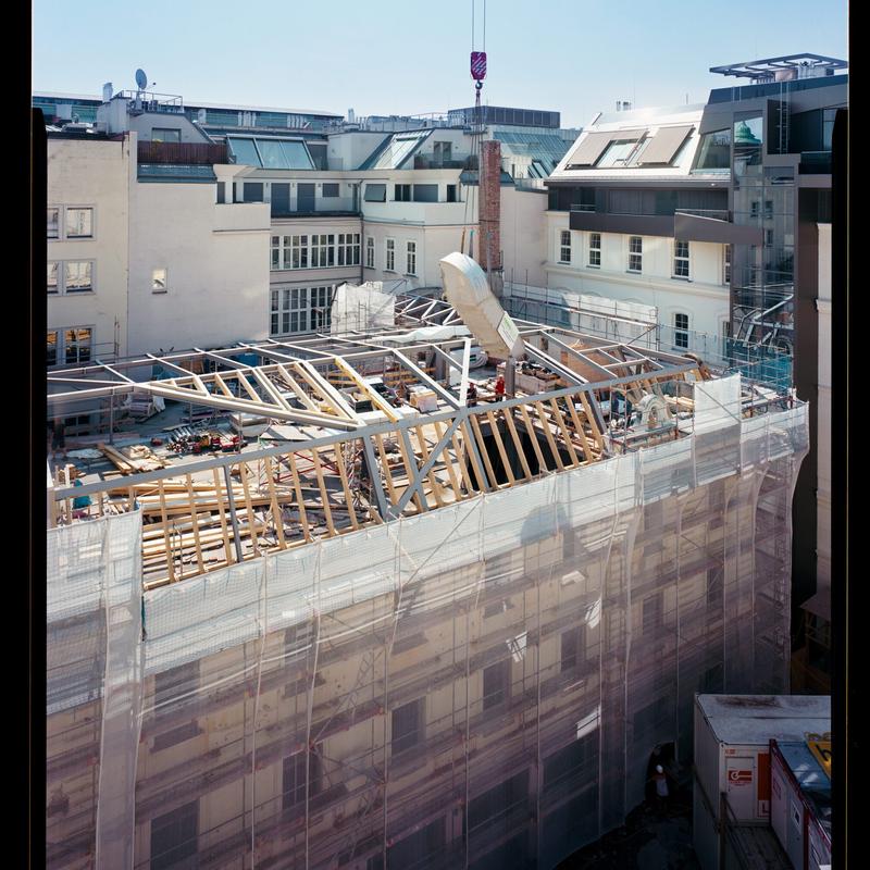 Blick auf eine Baustelle mit freiliegendem Dachstuhl, umgeben von Gerüst und Baucontainern, bei klarem Himmel.