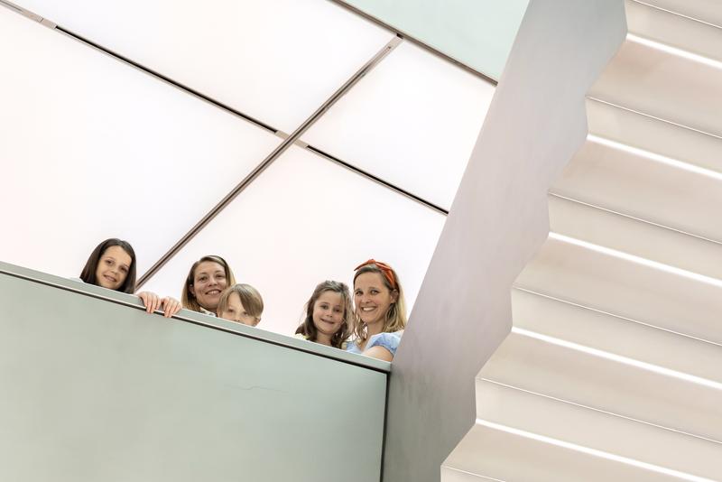 Auf dem Foto sieht man fünf Personen, darunter drei Kinder und zwei Erwachsene. Sie stehen auf einer Ausstellungsebene  und schauen freundlich in die Kamera. Alle lächeln.

Rechts im Bild sieht man eine moderne, helle Treppe.  Hinter den Personen sieht man die weiße Lichterdecke.