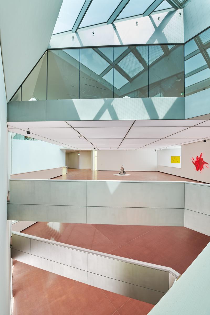 Moderne Architektur mit Glasdach, hellen Wänden und rotbraunem Boden, Skulptur und Kunstwerken im Raum.