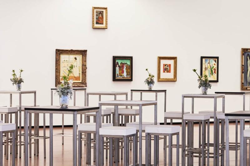 Moderne Galerie mit hohen Tischen, weißen Stühlen und mehreren gerahmten Kunstwerken an der Wand.