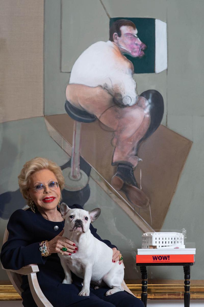 Auf dem Foto sieht man Heidi Horten.
Sie sitzt auf einem Sessel und hält einen kleinen weißen Hund auf dem Schoß.
Heidi Horten trägt dunkle Kleidung, große Schmuckstücke und eine blaue Brille.
Sie lächelt freundlich in die Kamera.

Hinter ihr hängt ein großes Gemälde von Francis Bacon.
Das Bild zeigt eine sitzende Figur, die sehr ausdrucksstark und ungewöhnlich gemalt ist – typisch für Bacons Stil.

Neben Heidi Horten steht ein kleiner Tisch.
Darauf liegen Kataloge, z.B. der "WOW" Katalog der Heidi Horten Collection.
Oben auf dem Tisch sieht man ein weißes Modell des Museumsgebäudes.