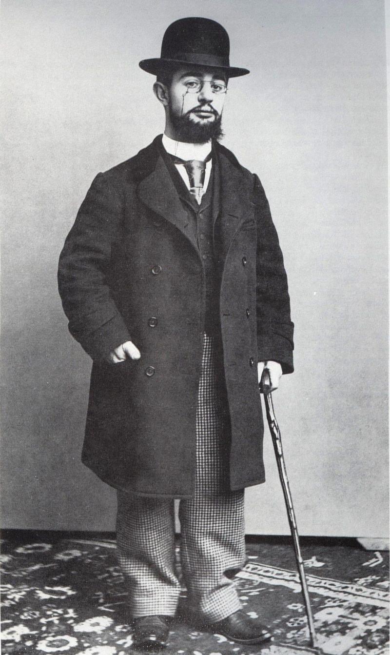 Das Foto zeigt den Künstler Henri de Toulouse-Lautrec. Er steht auf einem gemusterten Teppich vor einer schlichten Wand. Toulouse-Lautrec trägt einen dunklen Mantel, eine kariert gemusterte Hose und einen Hut. Er hat einen Schnurrbart und Bart und blickt ruhig in die Kamera. In seiner rechten Hand hält er einen Spazierstock, seine linke Hand steckt locker in der Manteltasche. Henri de Toulouse-Lautrec war ein französischer Maler und Plakatkünstler und ist bekannt für seine Darstellungen des Pariser Nachtlebens im 19. Jahrhundert. Er war kleinwüchsig und hatte durch eine Erkrankung Schwierigkeiten beim Gehen – darum nutzte er oft einen Stock zur Unterstützung.