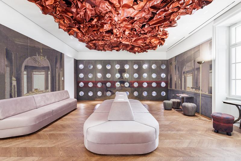 Auf dem Foto sehen wir den Tea Room im Museum der Heidi Horten Collection in Wien.

An der Decke hängt ein großes, rotes Kunstwerk von Hans Kupelwieser.
Es sieht aus wie zerknittertes Papier oder glänzender Stoff,
wurde aber aus Aluminium gemacht.

An der hinteren Wand gibt es viele runde Fenster – sie sehen aus wie Bullaugen.
Dahinter befinden sind kleine, kostbare Objekte aus der Sammlung von Heidi Horten.

Diese Vitrinenwand wurde von Markus Schinwald entworfen.
Auch die rosa Sitzmöbel in der Mitte des Raums gehören zu seinem Konzept.
Sie laden ein, sich hinzusetzen, zu schauen und zu entspannen.

Die Wände sind mit großen Stoffbildern bedeckt,
die an ein altes Café oder Schlosszimmer erinnern.