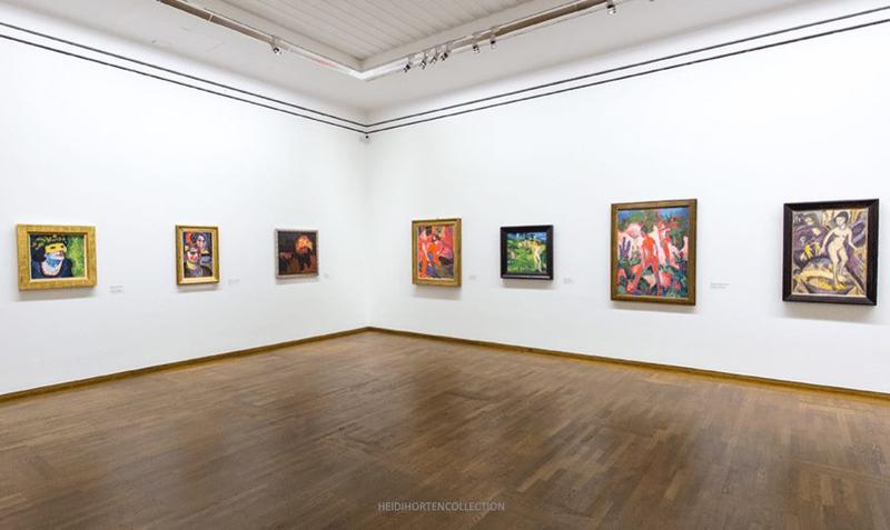 Ausstellungsraum mit mehreren Gemälden an weißen Wänden und einem Holzboden.